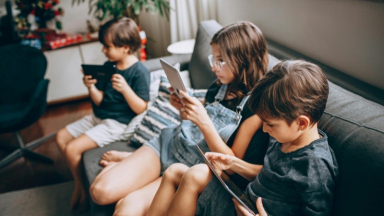 Tres niños sentados en un sofá, cada uno absorto en su propia tableta o consola portátil
