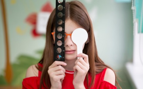 Niña con gafas se tapa un ojo con un oclusor mientras sostiene una regleta de lentes durante un examen de salud visual.